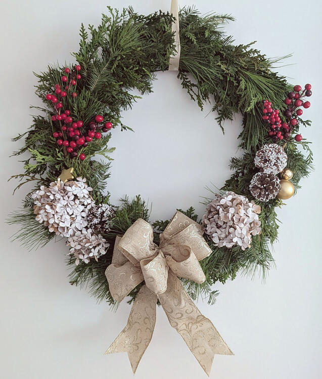 Christmas wreath with fresh evergreen foliage, dried hydrangea flowers and pine cones, artificial berries, and a gold bow