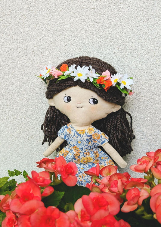 a fabric doll with an embroidered face, dark brown yarn decorated with a flower crown, in a blue-orange dress, stands in front of a white wall behind red begonias